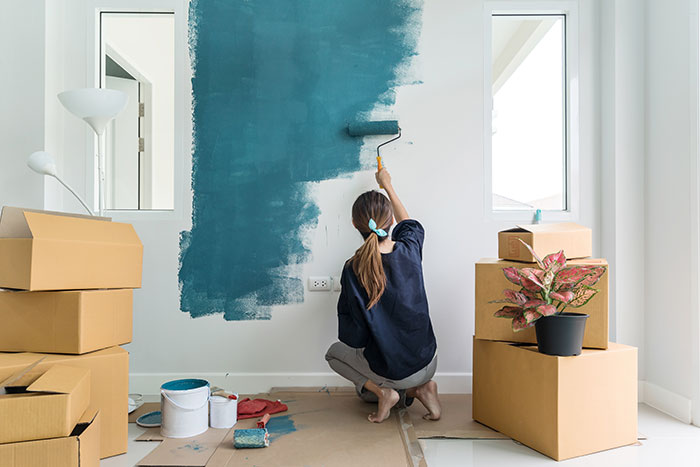 Woman painting a white wall in her home teal. She is surrounded by moving boxes.